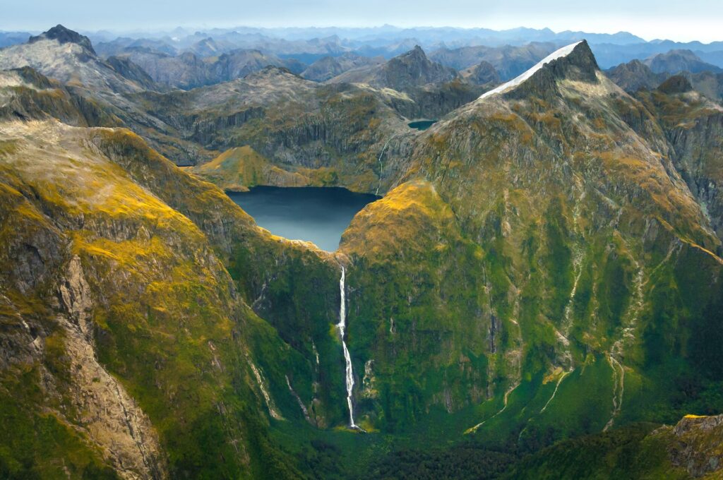 Green and yellow covered peaks tower around a bright blue lake with a thin, white waterfall cascading downwards at Milford Sound in New Zealand.