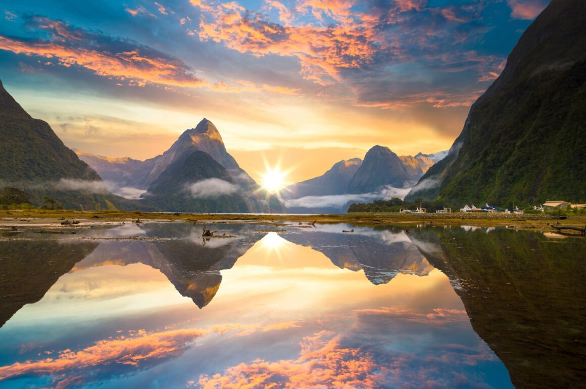 Triangle peaks are reflected in the water at Milford sound at sunset, with a dramatic yellow, blue and orange sky.