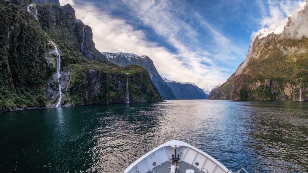 The bow of a boat on dark blue waters surrounded by mountains and waterfalls.