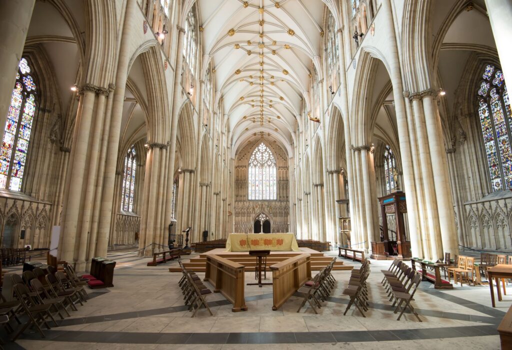 The interior of York Minster shows ornate ceilings in white marble, stained glass windows and pews and wooden chairs.