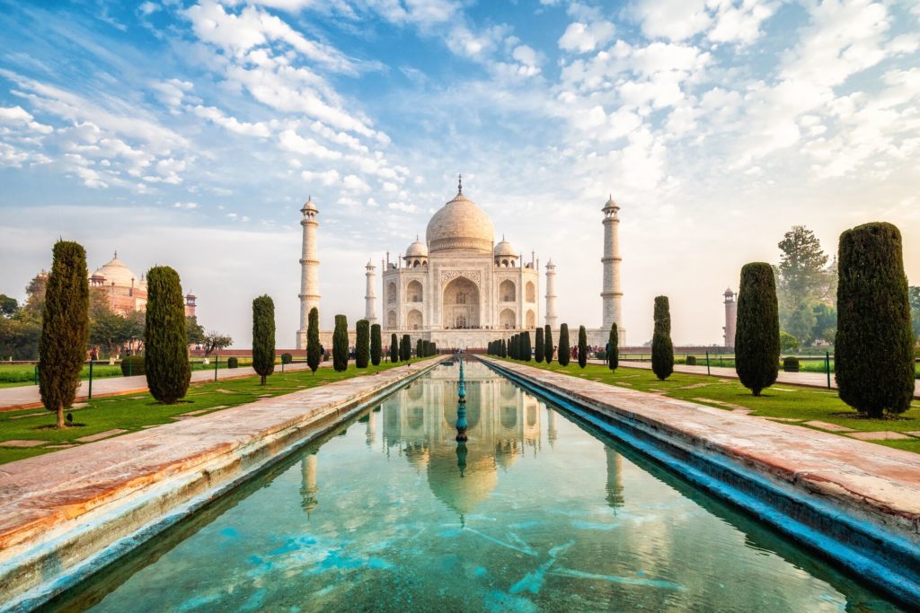 The Taj Mahal in luxury destination India stands at the end of a blue pond with ornate trees lining the sides. 