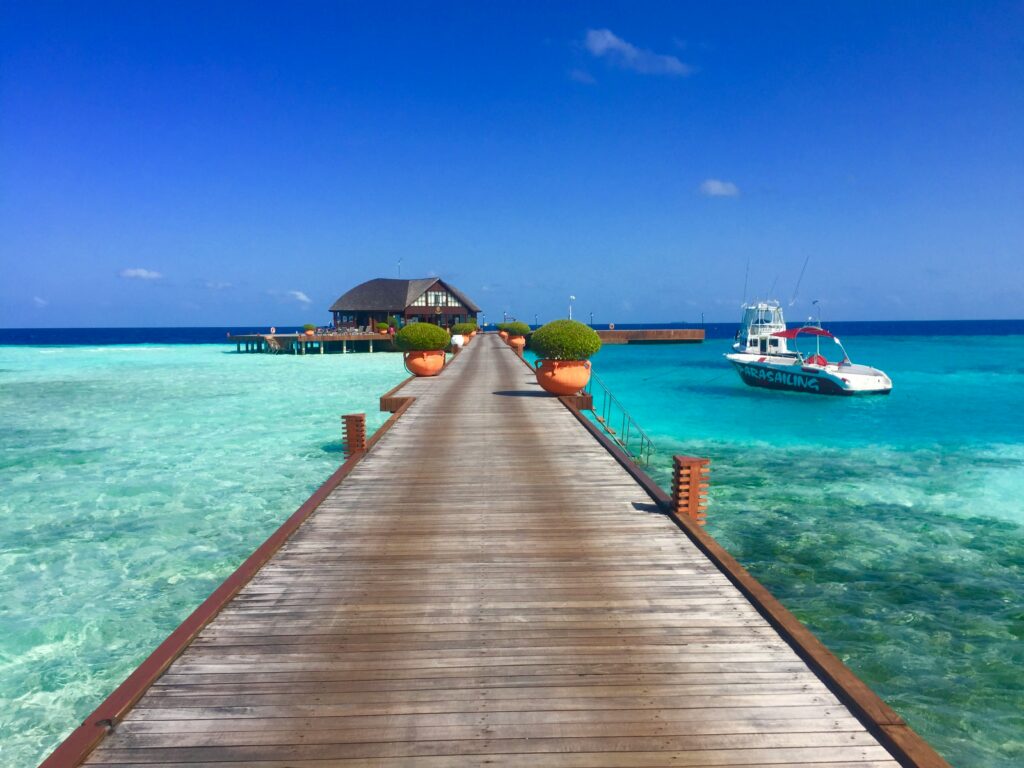 a wooden bridge stretches to a private over water villa with azure waters beneath and a bright blue sky.