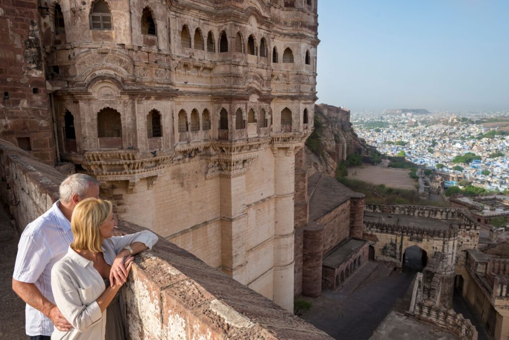 Guests look out from a bridge over the blue buildings in Jodhpur in India, with a large ornate tower beside them.