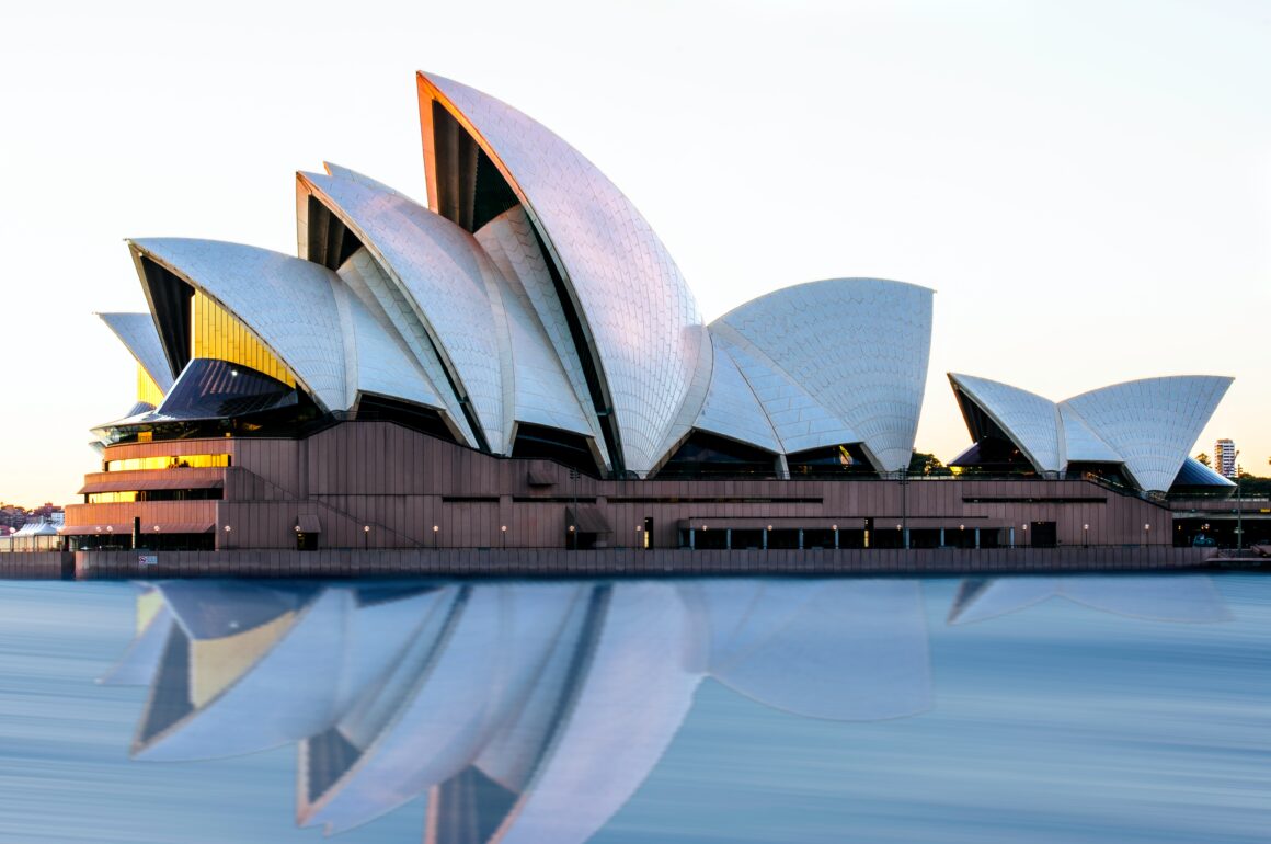 Sydney Opera House in the evening sun, with a full rsilver shining eflection of its dramatic architecture in the blue harbour waters below