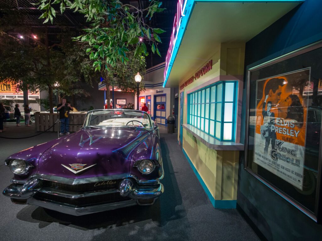 A purple car stands outside the Graceland car museum, home of Elvis preset, with a building to its side with blue neon window lights