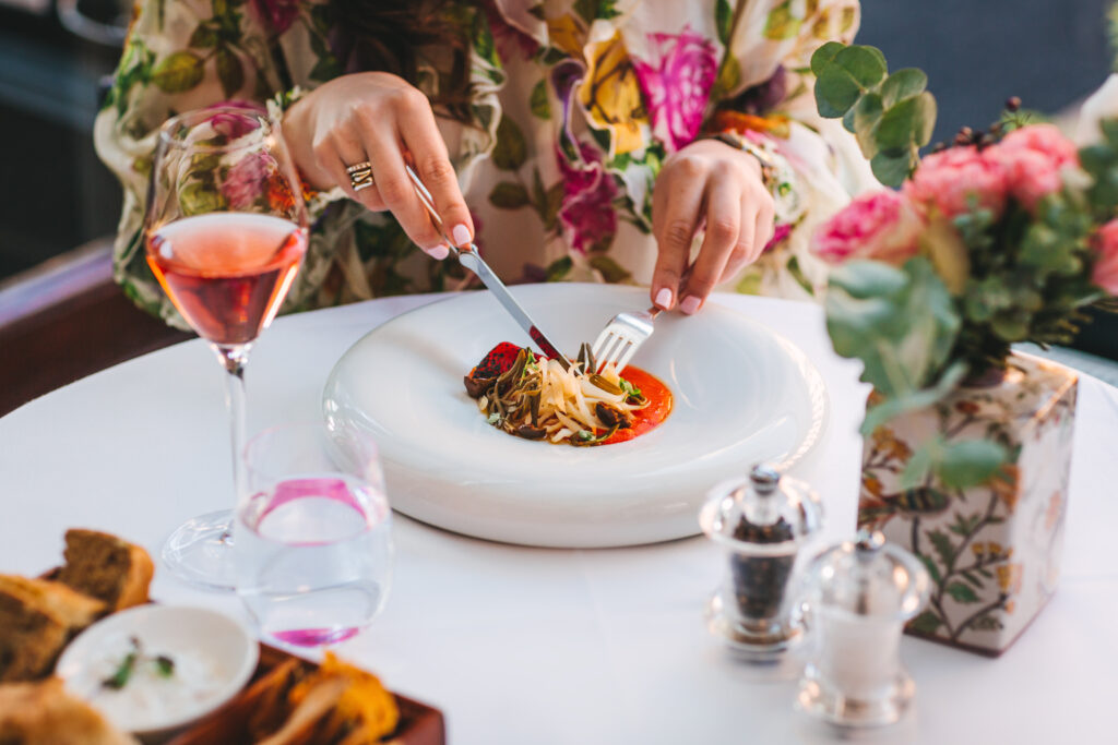 A woman in floral outfit cuts into a beautifully plaid dish, set on a table with a wine glass and flowers