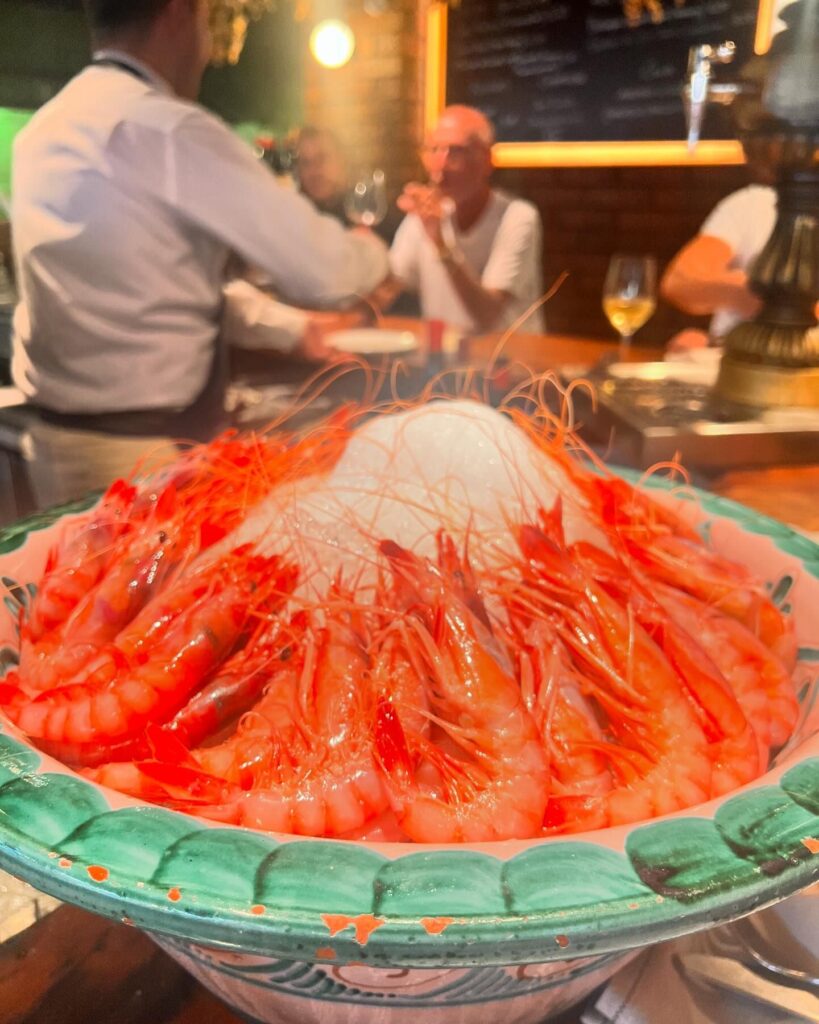 Close up of dish of bright red shrimp arranged around a white, cone shaped base on a bright green plate, with people dining in the background