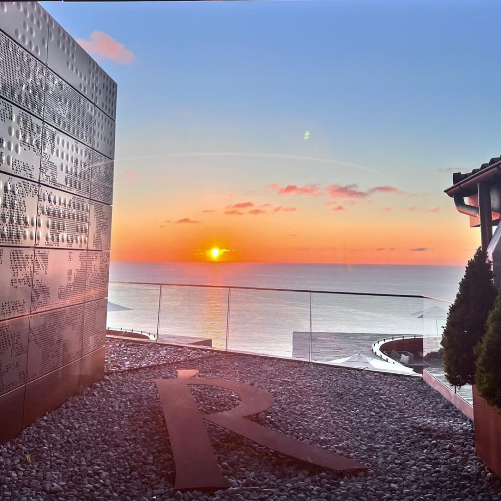 The sun sets on an ocean view, seen from a brown, iron clad building with a large metal A in the foreground, a restaurant for fine dining in Spain