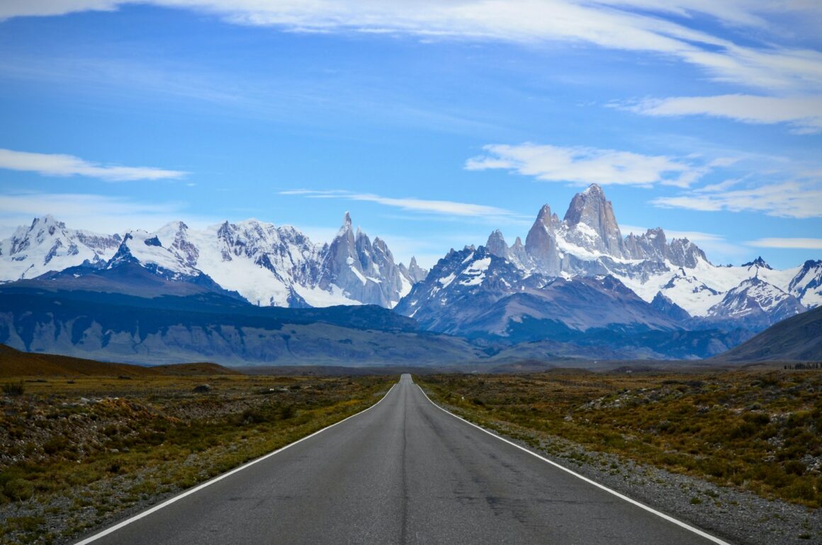 View looking down a road towards a jagged mountain range. Blue sky with a light streaking of cloud