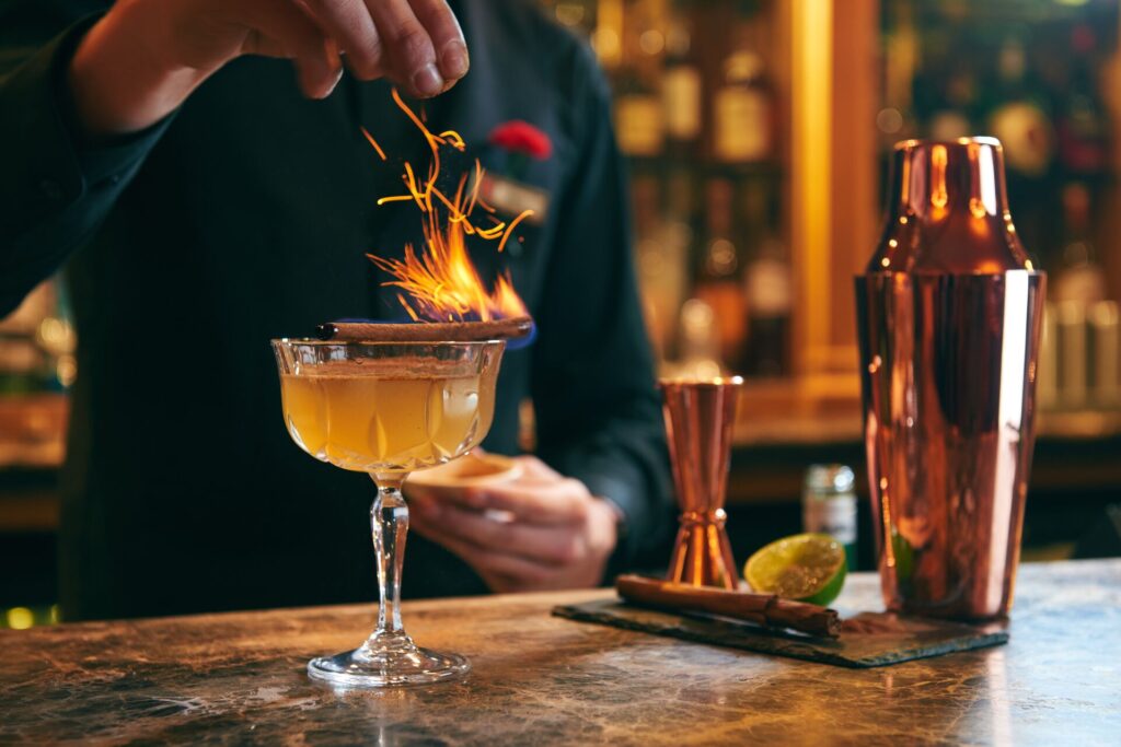 A cinnamon stick burns brightly on top of a yellow cocktail, with the mixologist standing behind.