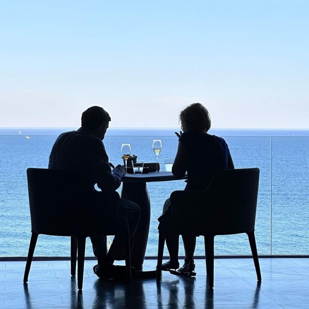 A couple dine overlooking he blue ocean, shown in black shilouette