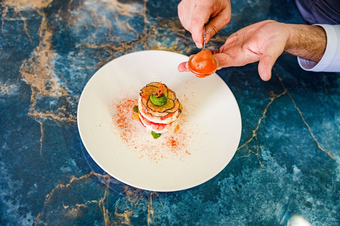 Tow hands prepare to place a tomato onto a delicate colourful stack of food on a white dish, on a blue marble tabletop