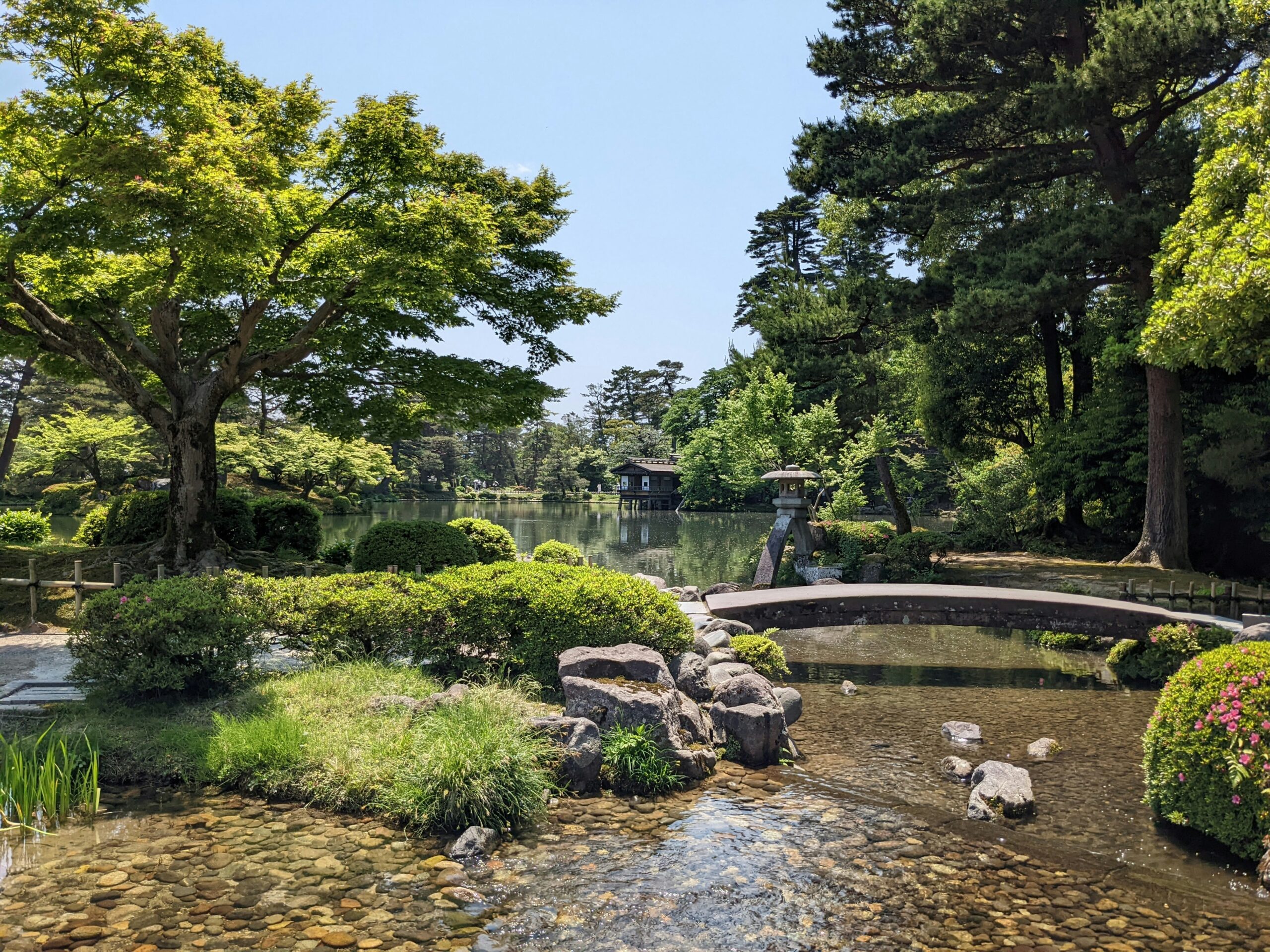 Kenrokuen Garden, Kanazawa in Japan with a low bridge over wise waterways, landscaped green grass and trees and a bright blue sky