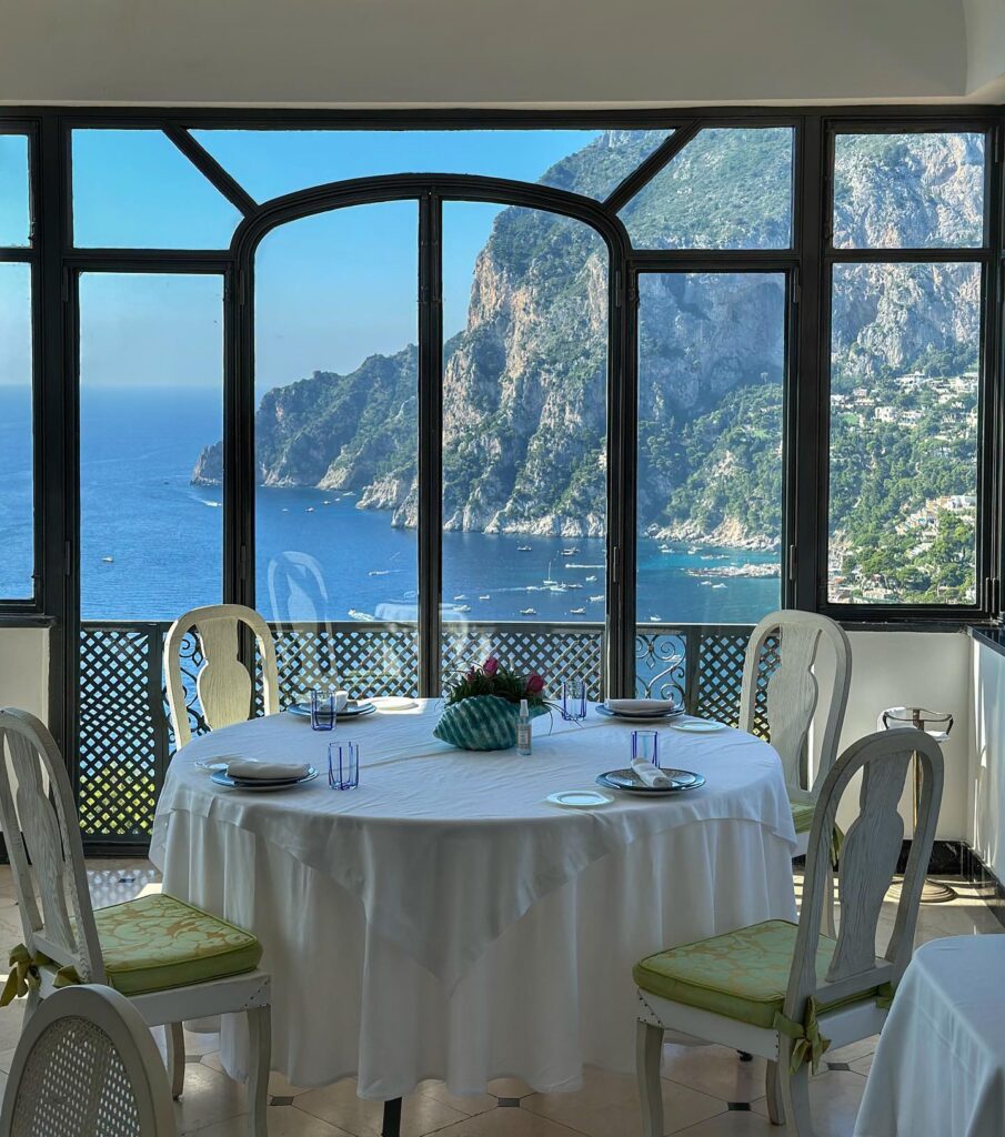 A round table with white linen is set for dinner in front of a window showing a view of mountains and sea in Italy