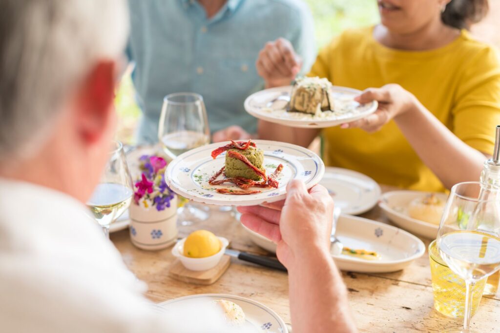 People dine around a wooden table with china dishes with elegant plates of rustic food.