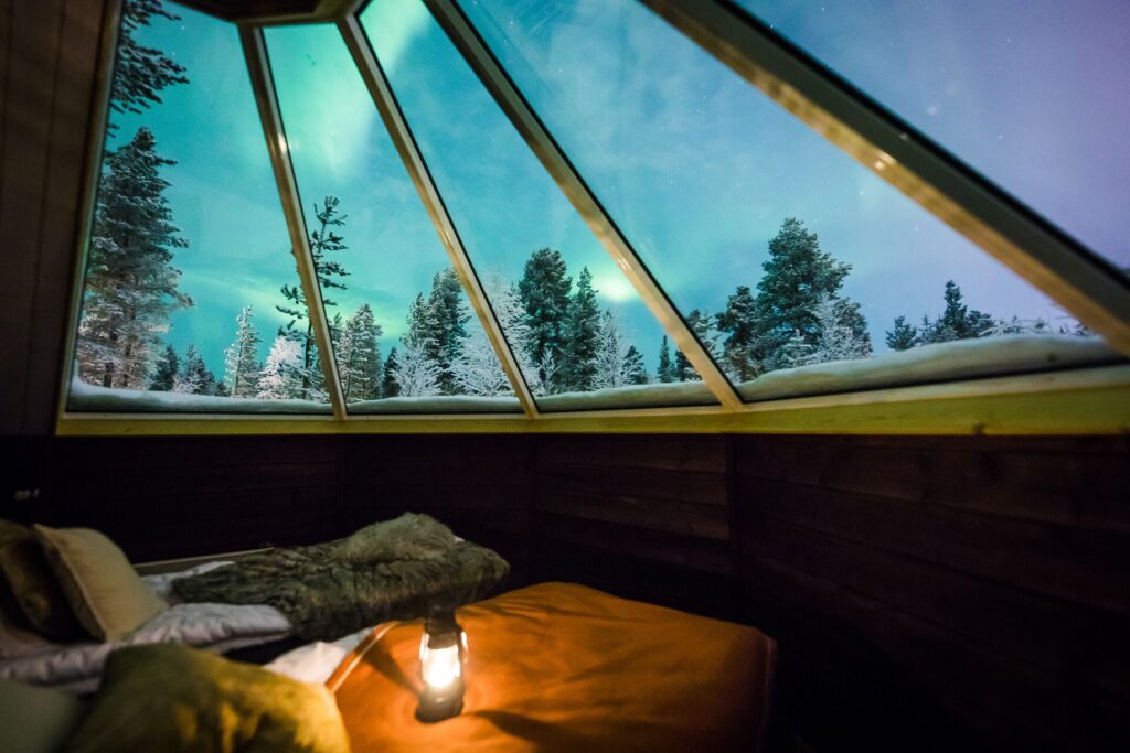 The arctic sky at night with blue and green Northern Lights is seen through a glass panelled hotel roof with a bed below.