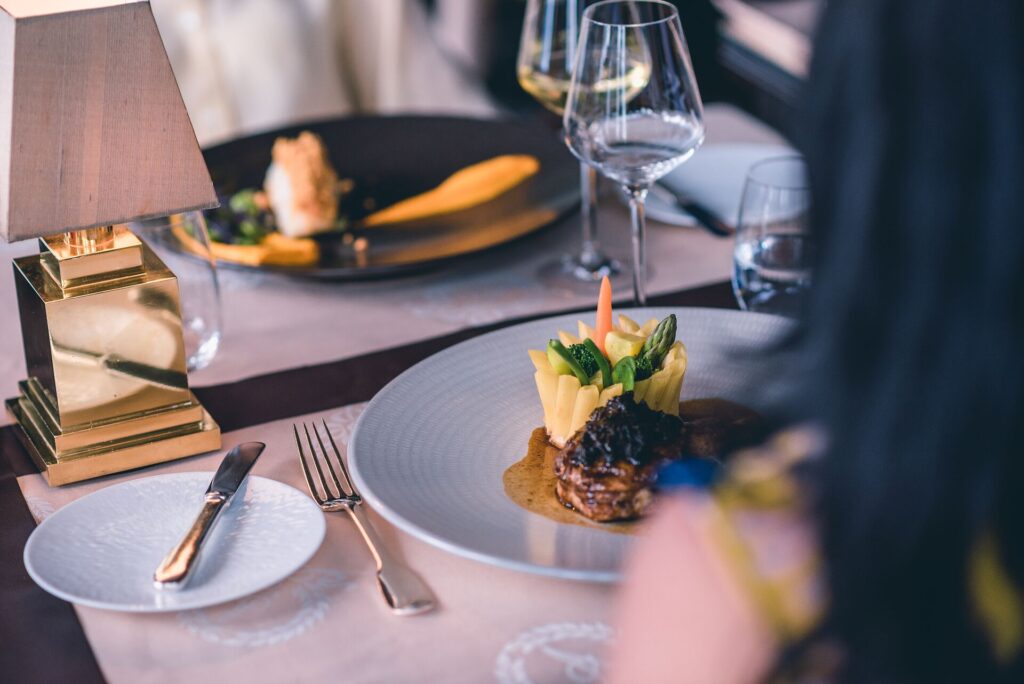 An elegant plat of food, artistically presented on a white plate on a laid table with wine glasses, cutlery and a lamp.