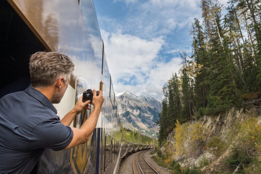 Man leaning out of a train with a camera taking a photo of the mountains ahead