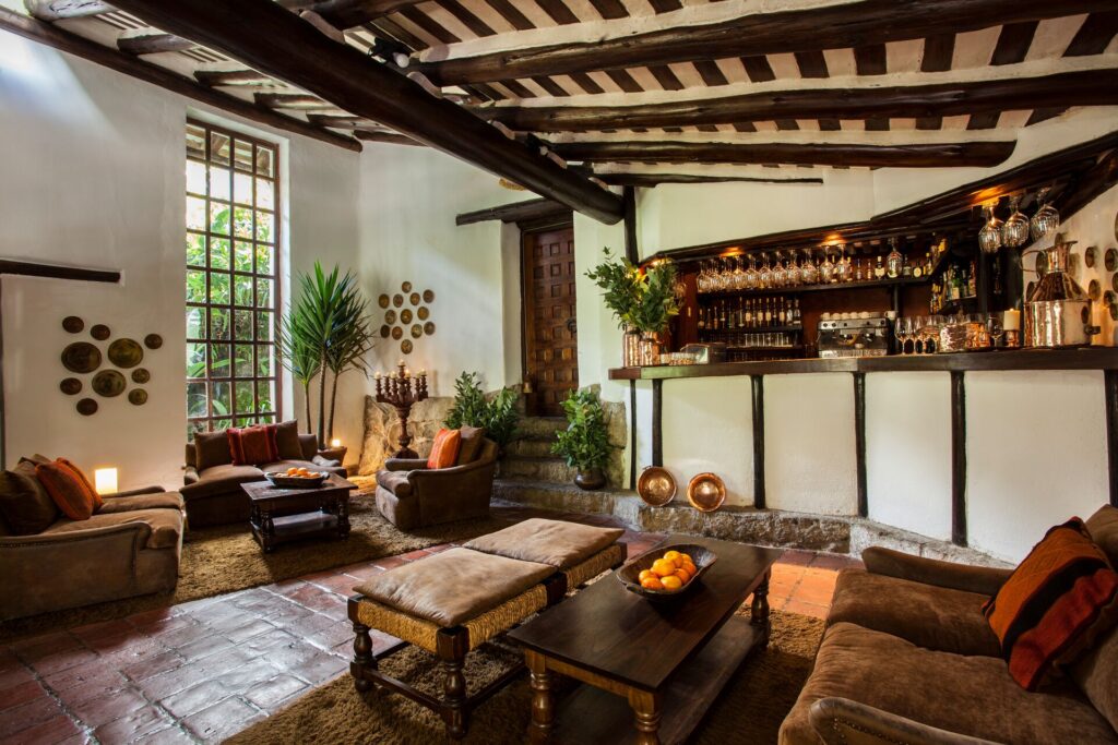 A traditional Peruvian hotel room with white walls, dark wooden beams, plants and comfy brown furniture.