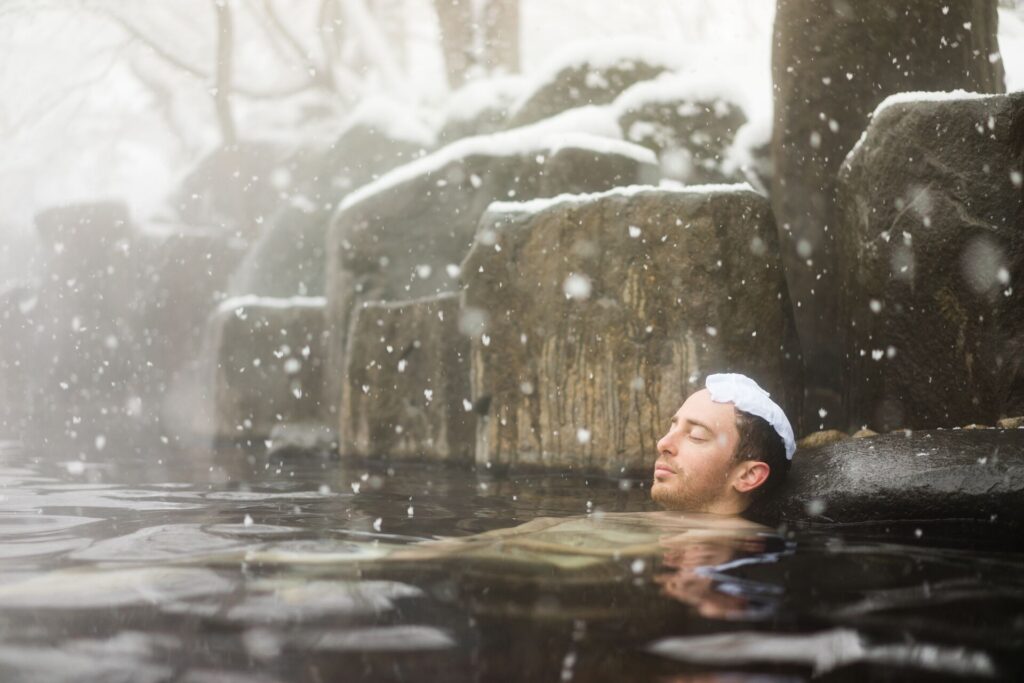 A mad relaxes up to his neck in water in a natural spring, with show flakes falling around him. 