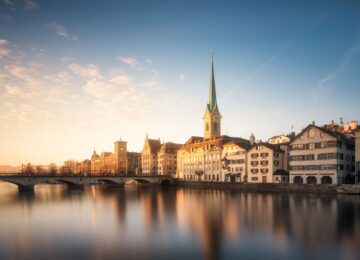 A tall emerald green church spire sits among a row of buildings, with a river an bridge in front of in Zurich, Switzerland.