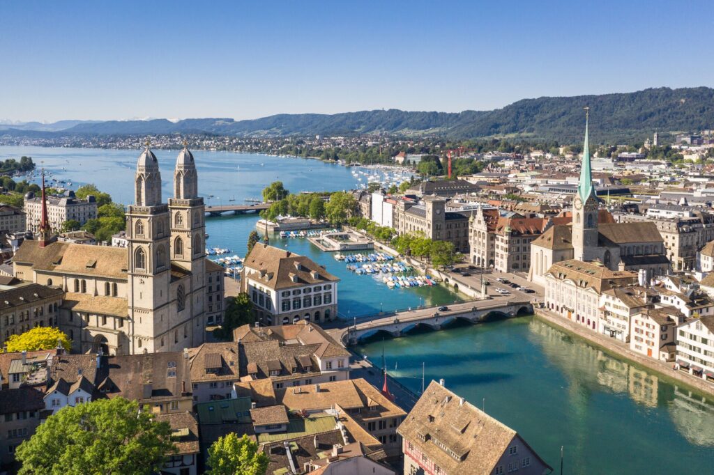 Aerial shot of Zurich, with buildings set alongside a bright blue lake, towering church spires and bridges over a river.