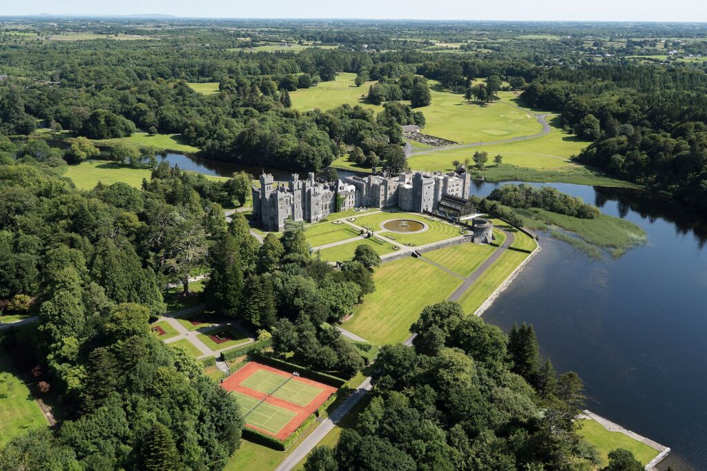 Aerial shot of Ashford Castle in Ireland, an enormous light grey fortress set among green lawns and woodland, with a blue lake in front.