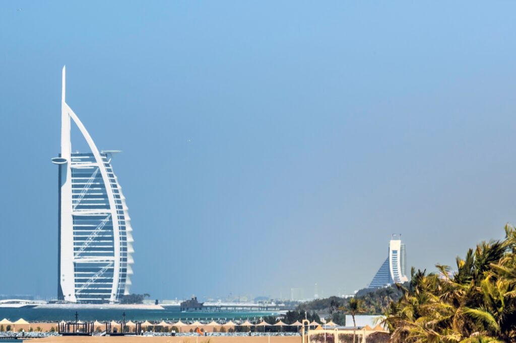 The Iconic white Burj Al Arab hotel in Dubai with a blue sky and beach in front lined with parasols.