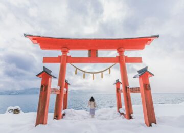 Woman photographed from behind standing beneath torii gate in snow overlooking lake