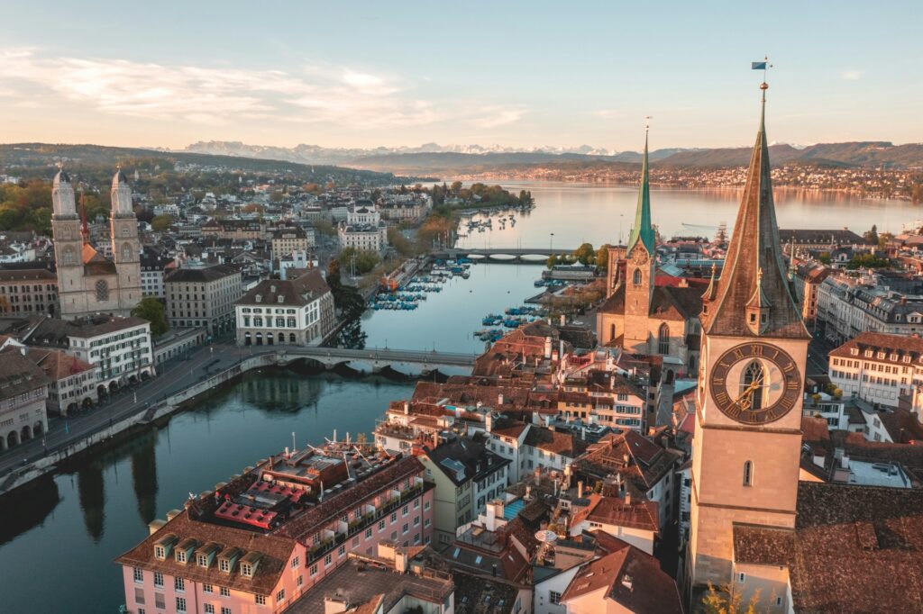 Aerial view of European city skyline, with church steeple in the foreground, lake and mountains in the background